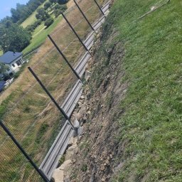 Piotr Salachna - Długi, panelowy płot z siatki, biegnący wzdłuż stromego zbocza z widokiem na dom w oddali i zielone wzgórza pod błękitnym niebem.