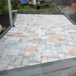 Świeżo ułożona, wielobarwna kostka brukowa tworzy geometryczny wzór na podjeździe. Szara obwódka podkreśla krawędzie, a w tle widoczny ogród i meble ogrodowe.