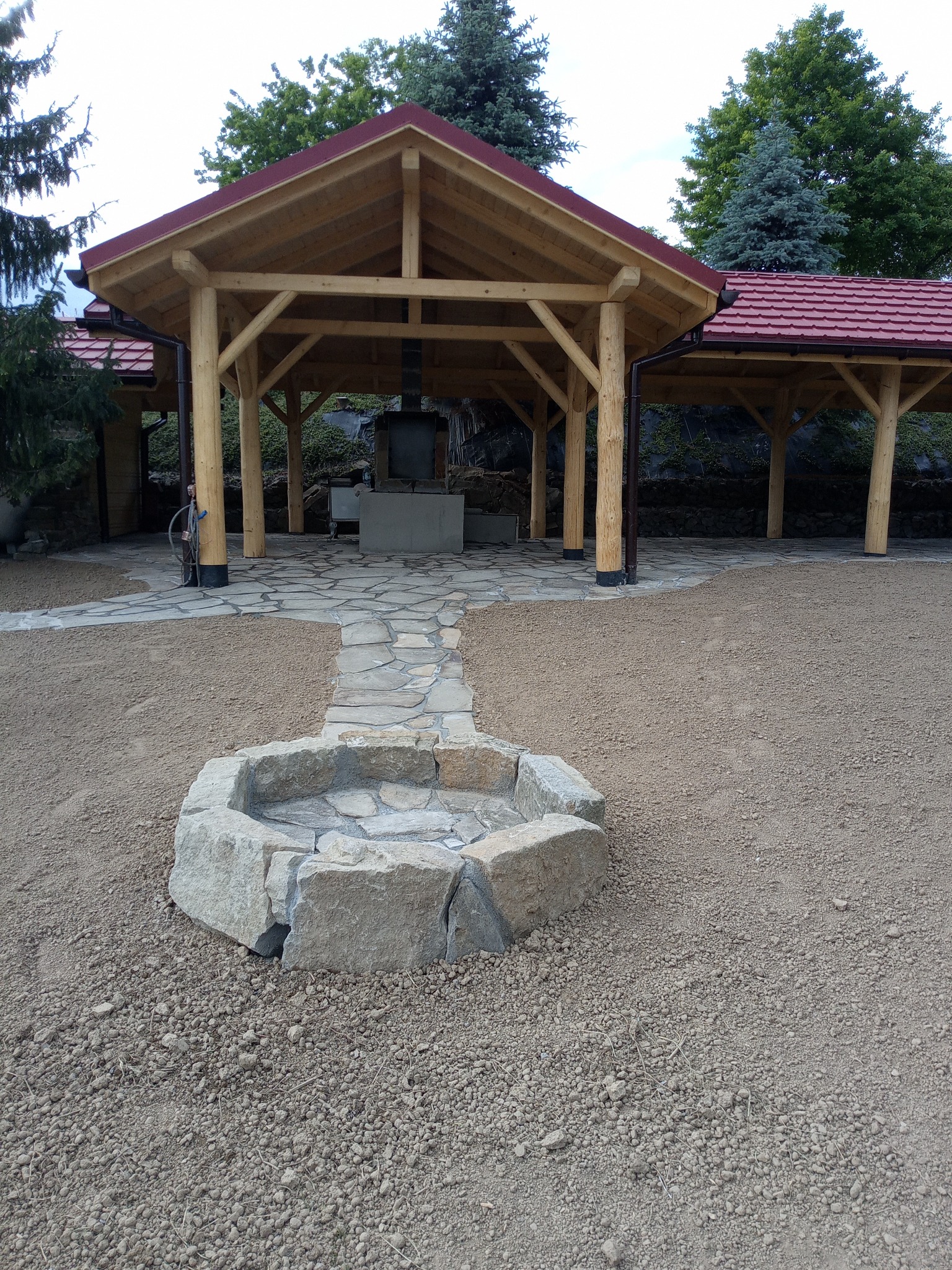 Zadaszona drewniana konstrukcja z czerwonym dachem, kamienny chodnik i okrągłe palenisko z kamienia na żwirowym podłożu.
