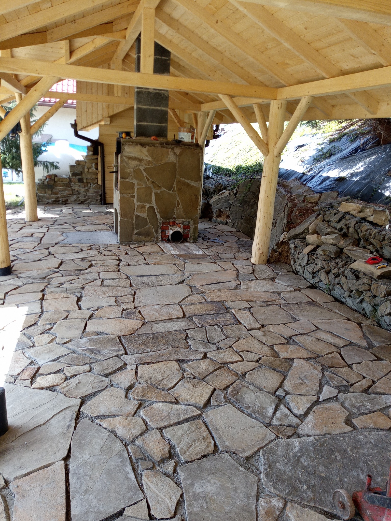 Zadaszona przestrzeń z nieregularną kamienną posadzką, murowanym kominkiem z kamienia łupanego i drewnianą konstrukcją dachu, otoczona kamiennym murem oporowym.