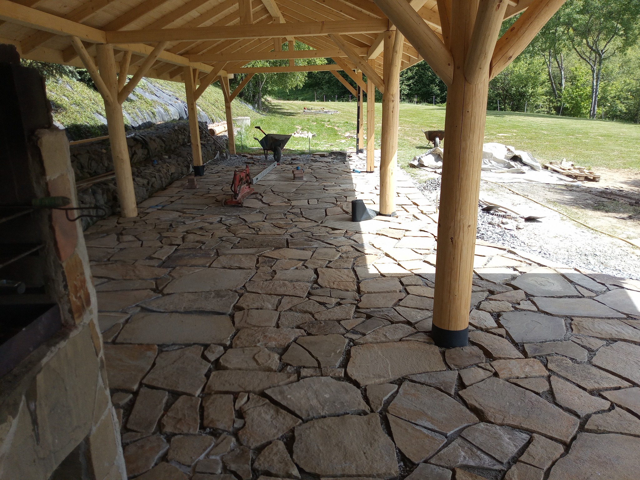 Nieregularna kamienna posadzka tarasu pod drewnianą konstrukcją z widocznymi narzędziami budowlanymi i taczką w tle.