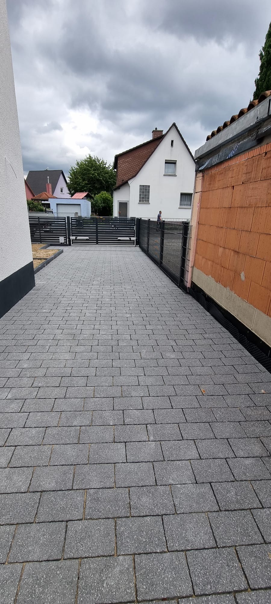 Ułożona szara kostka brukowa na podjeździe, z widocznym ogrodzeniem panelowym i fragmentem budynku w trakcie budowy w tle.