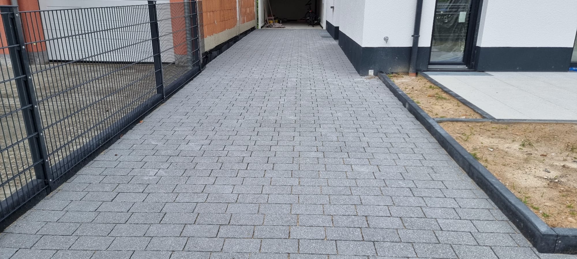 Ułożona szara kostka brukowa prowadząca do garażu, z widocznym ogrodzeniem panelowym po lewej stronie i fragmentem domu po prawej. Widoczny fragment nowej nawierzchni z jasnych płyt.