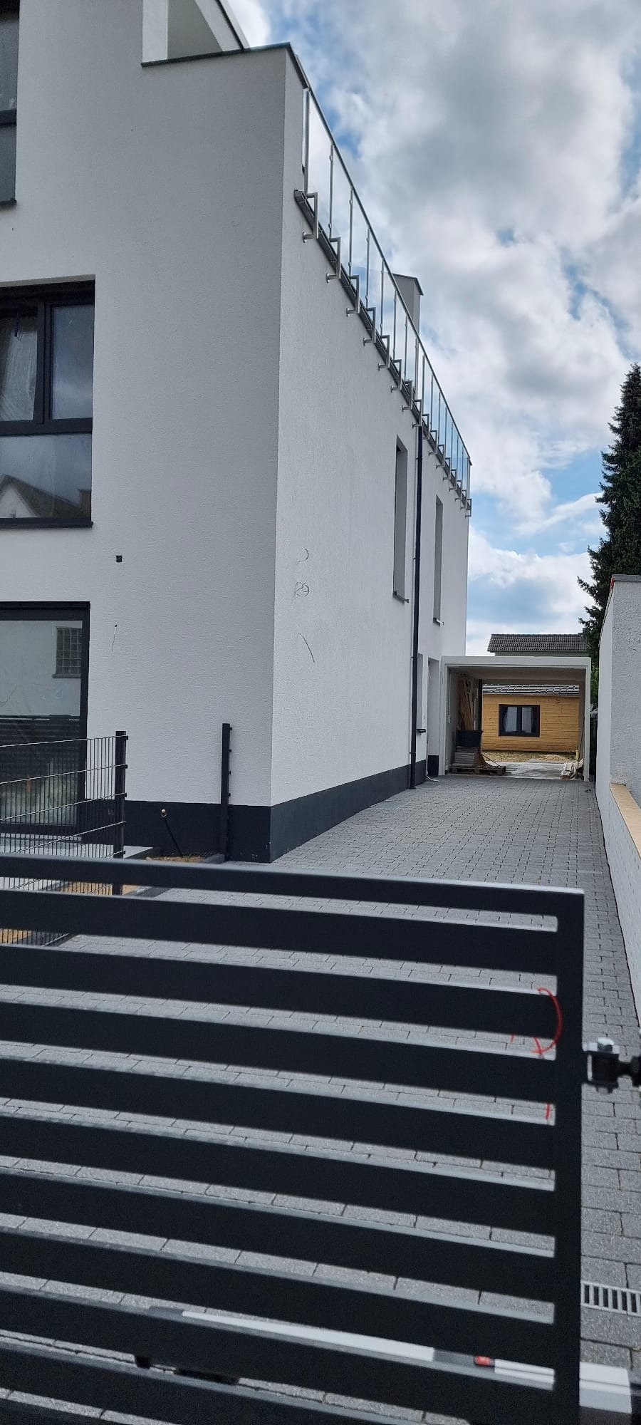 Nowoczesny dom z białymi ścianami, szarą kostką brukową na podjeździe i czarnym ogrodzeniem panelowym na pierwszym planie.