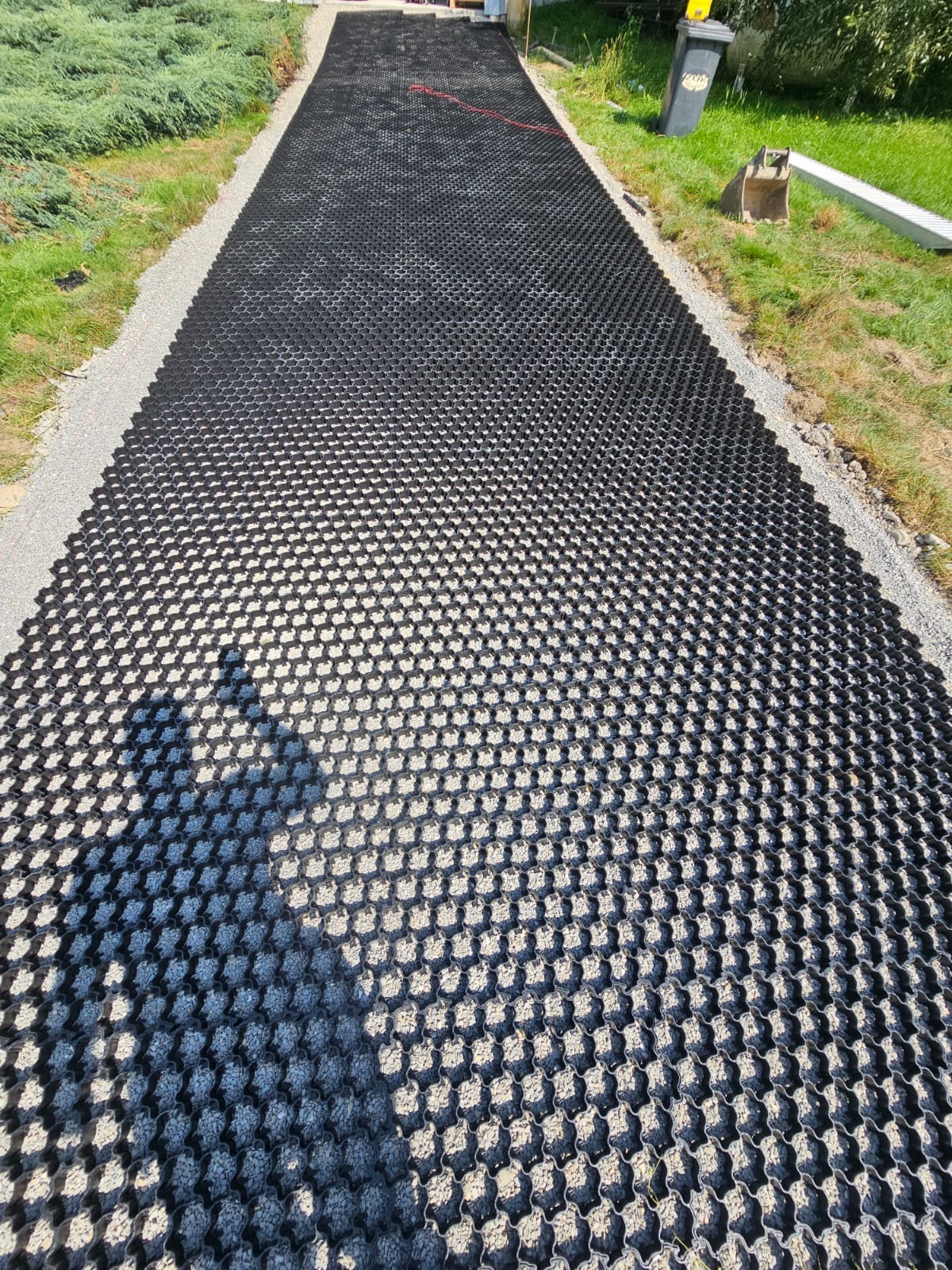 Długa droga z czarnych, ażurowych płyt z wypełnieniem z szarego kruszywa, obrzeża z kamyczków, trawa po bokach, widoczny cień osoby robiącej zdjęcie.