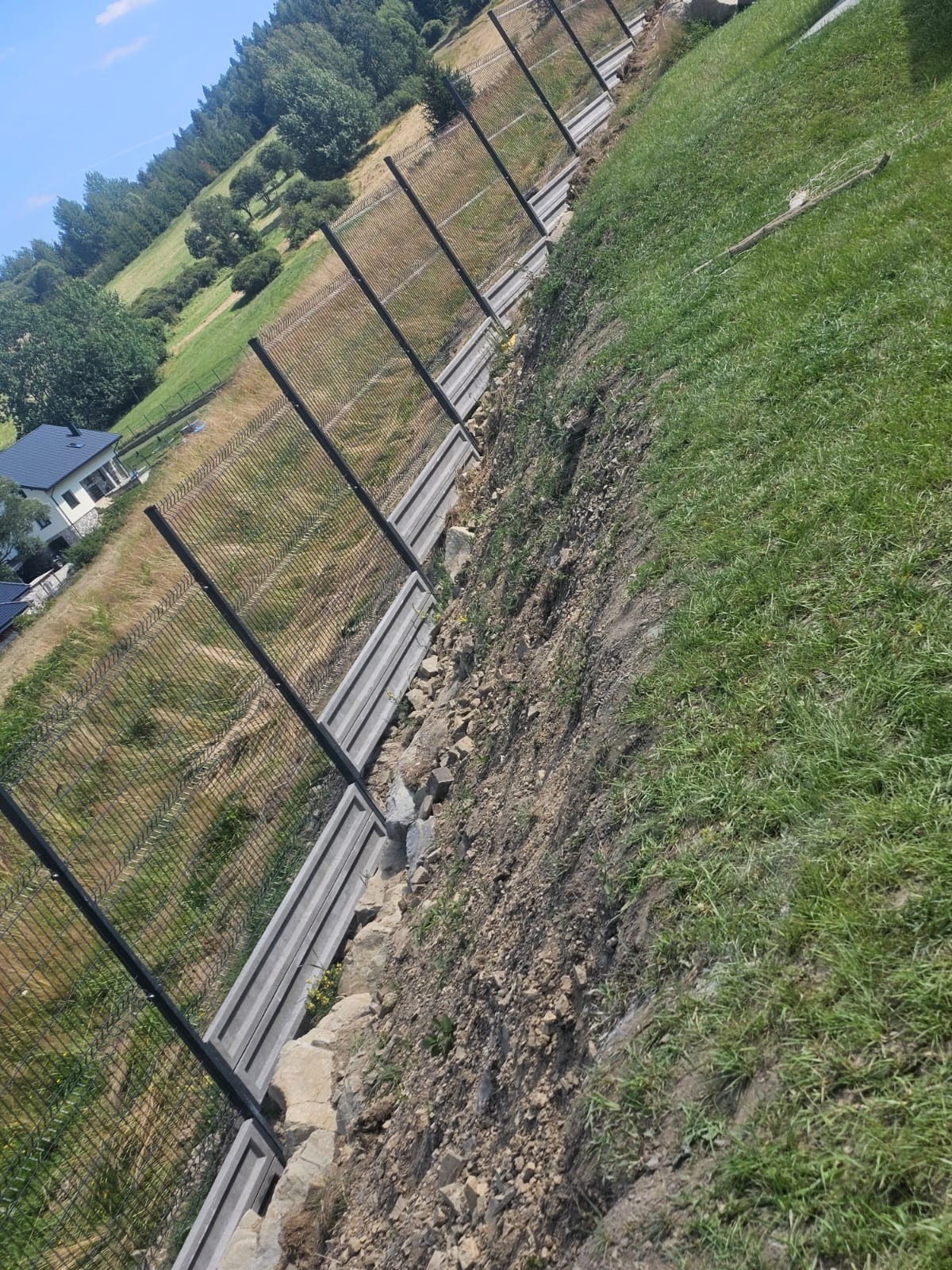 Długi, panelowy płot z siatki, biegnący wzdłuż stromego zbocza z widokiem na dom w oddali i zielone wzgórza pod błękitnym niebem.