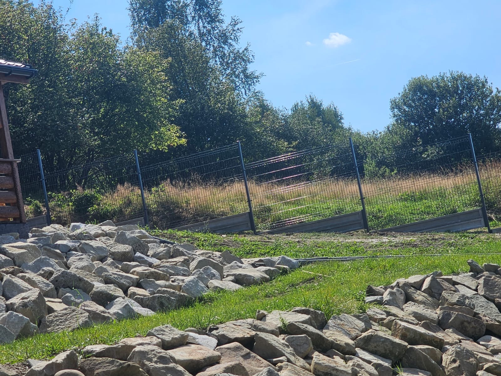 Metalowe ogrodzenie panelowe z podmurówką na zboczu, wzmocnione kamieniami, z widokiem na zieleń i błękitne niebo.