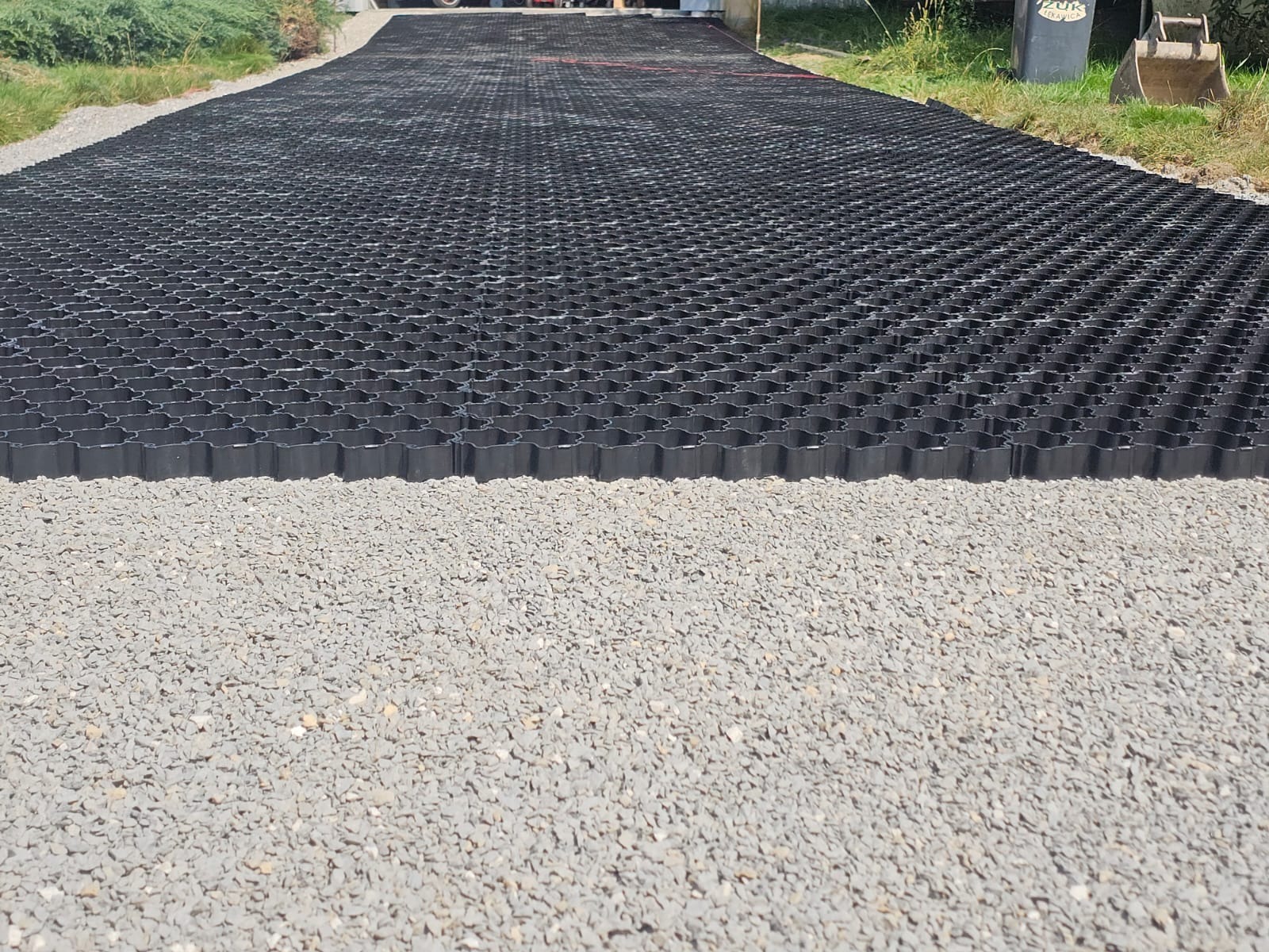 Czarna, plastikowa kratka trawnikowa ułożona na podłożu z drobnego szarego kruszywa, widok z perspektywy wzdłuż drogi, z fragmentem zieleni po bokach.