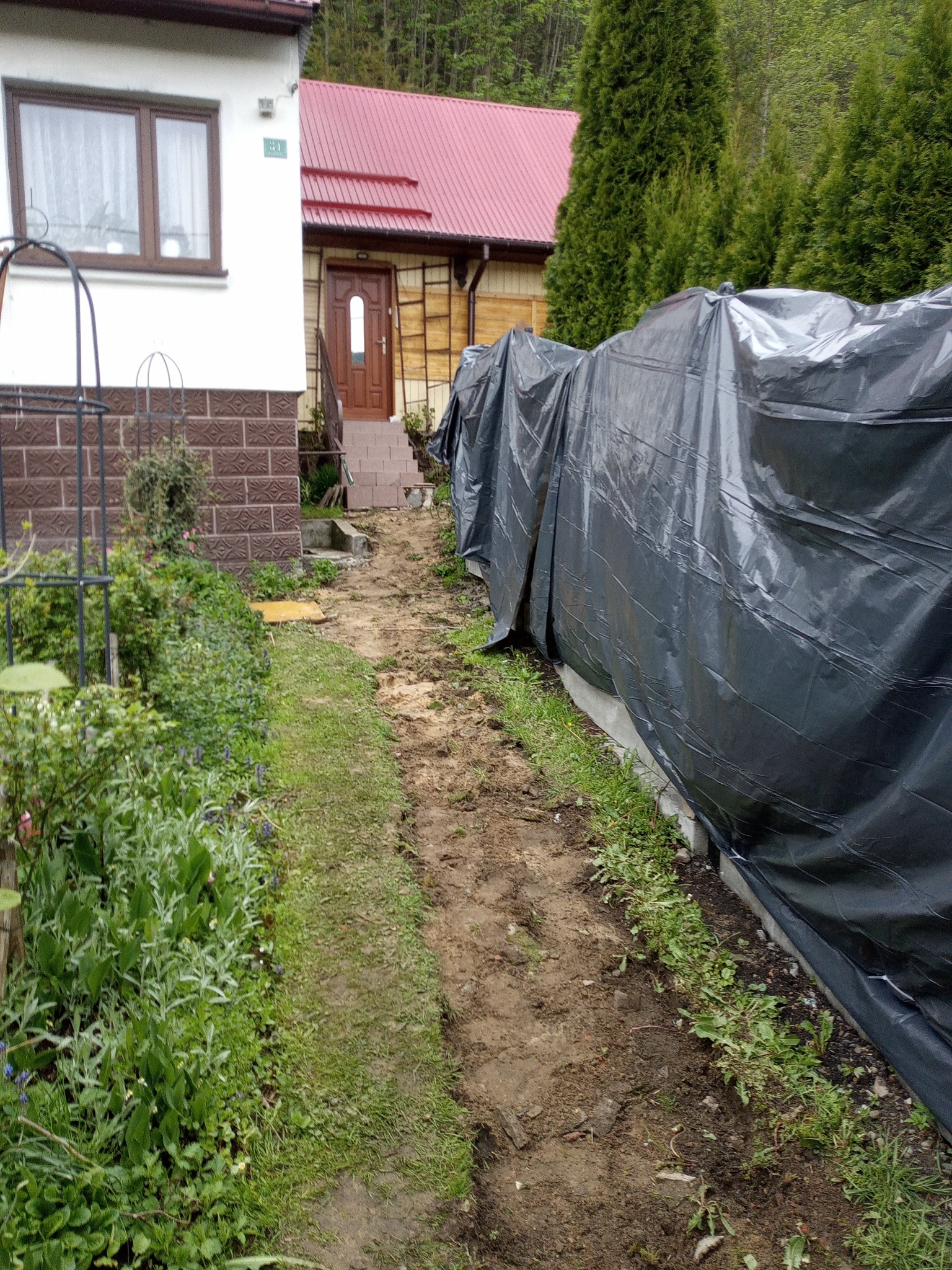 Przygotowanie terenu pod budowę ogrodzenia. Widoczna folia zabezpieczająca, wytyczona ścieżka i fragment domu z zielenią.