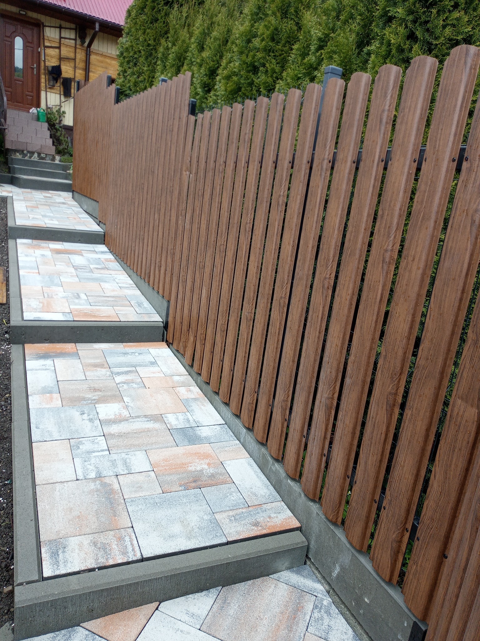 Schody z kostki brukowej prowadzące wzdłuż brązowego, panelowego ogrodzenia z pionowych desek, na tle zielonych krzewów i drewnianego domu.
