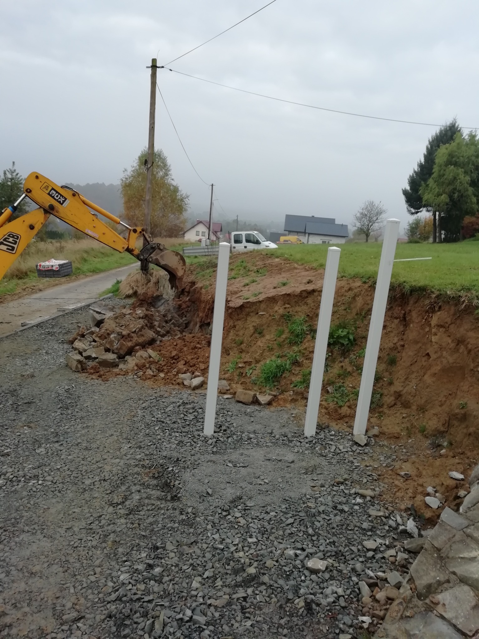 Żółta koparka przygotowuje teren pod montaż białych słupków ogrodzeniowych na żwirowej drodze. W tle domy i pochmurne niebo.