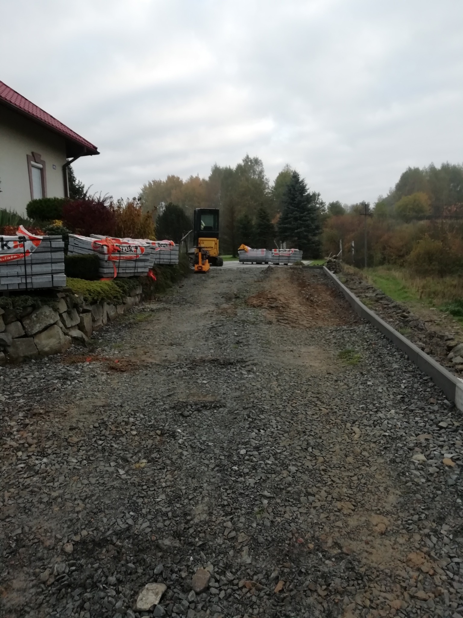 Przygotowanie podłoża pod kostkę brukową z mini koparką w tle. Ułożone obrzeża i palety z kostką. Widok na posesję i zieleń w tle.