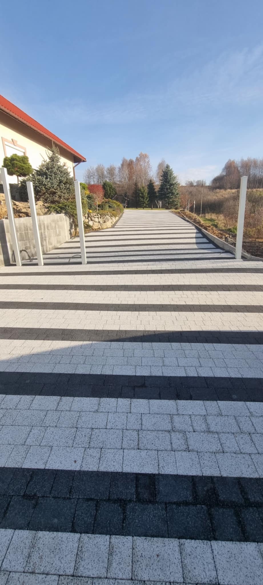 Szeroki podjazd z kostki brukowej w paski, prowadzący do domu z ogrodem. Białe słupki po bokach podjazdu. Widok z perspektywy.