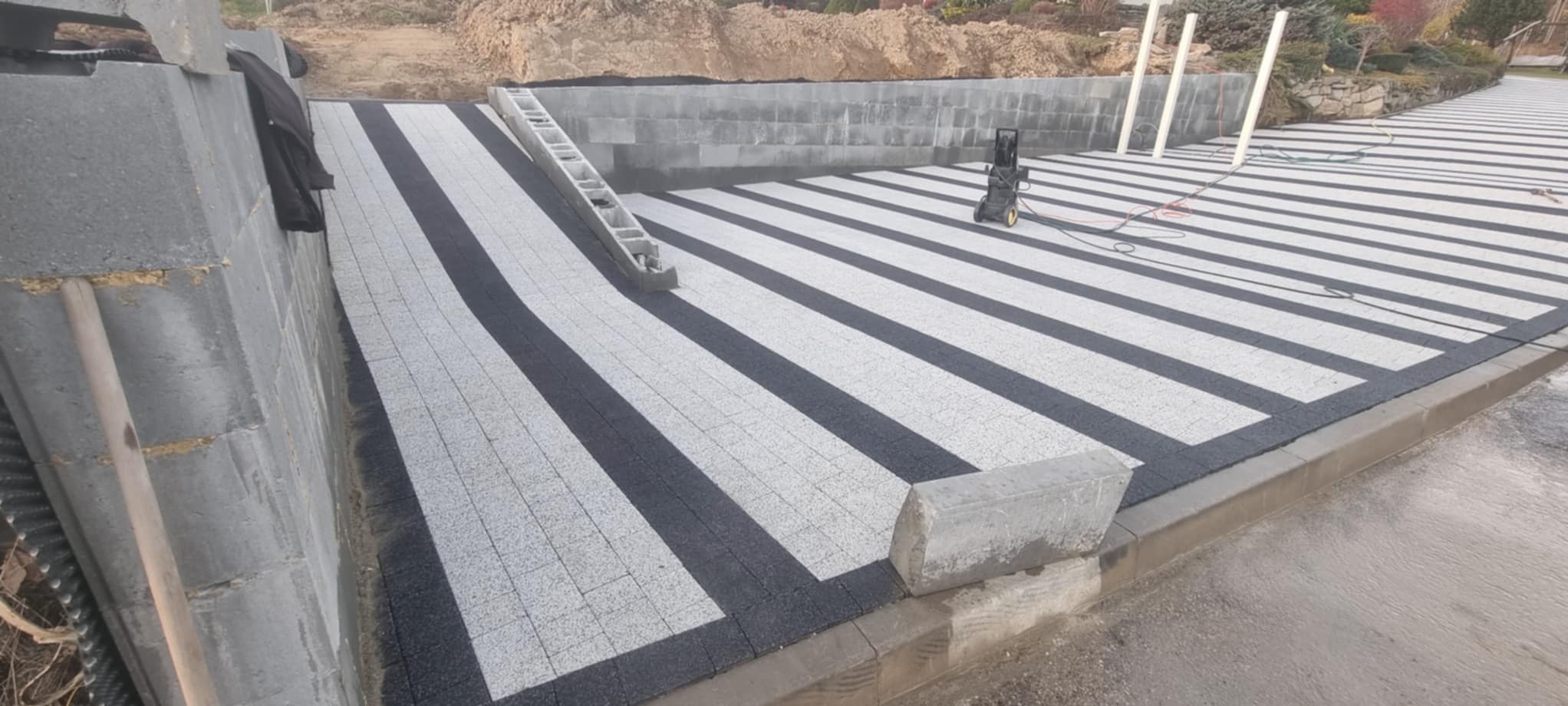 Ukończona nawierzchnia z kostki brukowej w biało-czarne pasy, z widoczną drabiną i myjką ciśnieniową. Świeżo ułożona kostka, widoczne elementy muru oporowego.