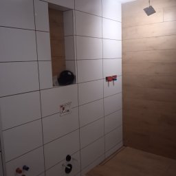 Artur Konieczny - Łazienka w trakcie wykańczania: białe płytki ścienne, drewnopodobne płytki podłogowe, przygotowane punkty instalacyjne, wnęka z czarnym kaskiem ochronnym.