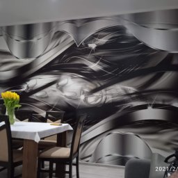 Artur Konieczny - Jadalnia z fototapetą w odcieniach szarości z motywem abstrakcyjnych, metalicznych fal i refleksów świetlnych. Na pierwszym planie stół nakryty białym obrusem z bukietem żółtych tulipanów.