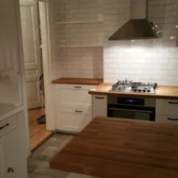 Kuchnia z białymi szafkami, drewnianym blatem i płytkami imitującymi cegłę na ścianie, widoczny fragment wyspy kuchennej z drewnianym blatem oraz otwarte drzwi do innego pomieszczenia z mopem...