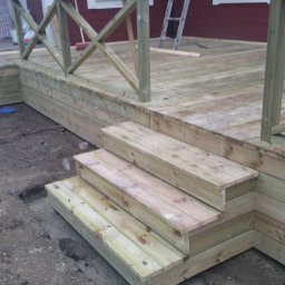 Artur Konieczny - Nowo wybudowany drewniany taras z balustradą i schodami, widoczny w surowym stanie przed wykończeniem, z ziemią wokół i fragmentem czerwonego budynku w tle.