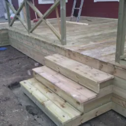 Nowo wybudowany drewniany taras z balustradą i schodami, widoczny w surowym stanie przed wykończeniem, z ziemią wokół i fragmentem czerwonego budynku w tle.