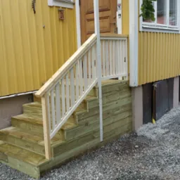 Nowe drewniane schody z białą balustradą prowadzące do żółtego domu z drewnianą elewacją i brązowymi drzwiami wejściowymi.