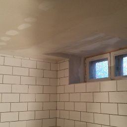 Artur Konieczny - Ściana wyłożona białymi, prostokątnymi płytkami z ciemną fugą, sufit w trakcie szpachlowania z widocznymi łatami gładzi, okno z ozdobną szybą w górnej części ściany.