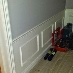 Artur Konieczny - Szara ściana z dekoracyjną białą boazerią panelową, narzędzia i skrzynki narzędziowe leżące na podłodze z paneli imitujących drewno, widok z dołu.