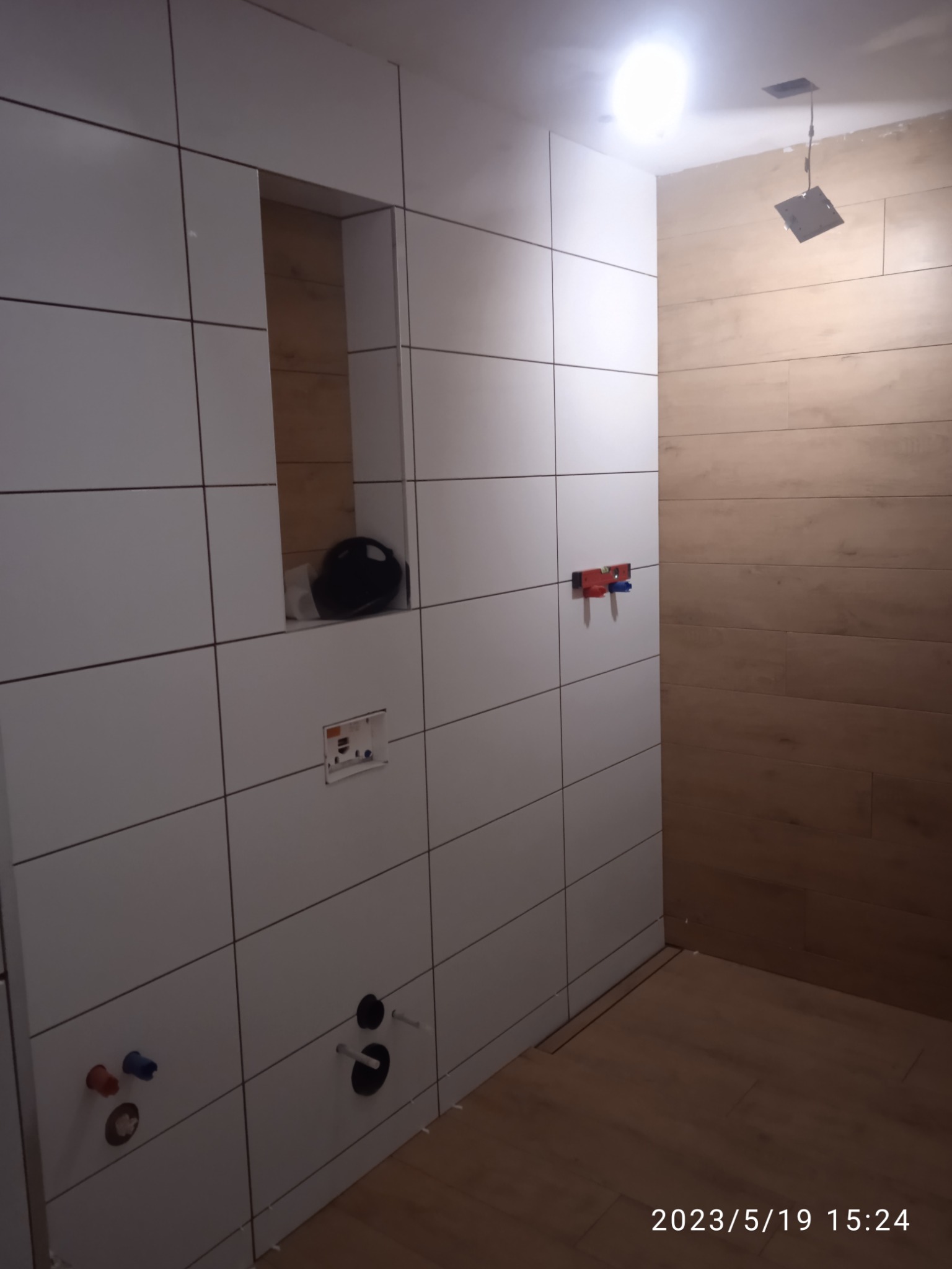 Łazienka w trakcie wykańczania: białe płytki ścienne, drewnopodobne płytki podłogowe, przygotowane punkty instalacyjne, wnęka z czarnym kaskiem ochronnym.