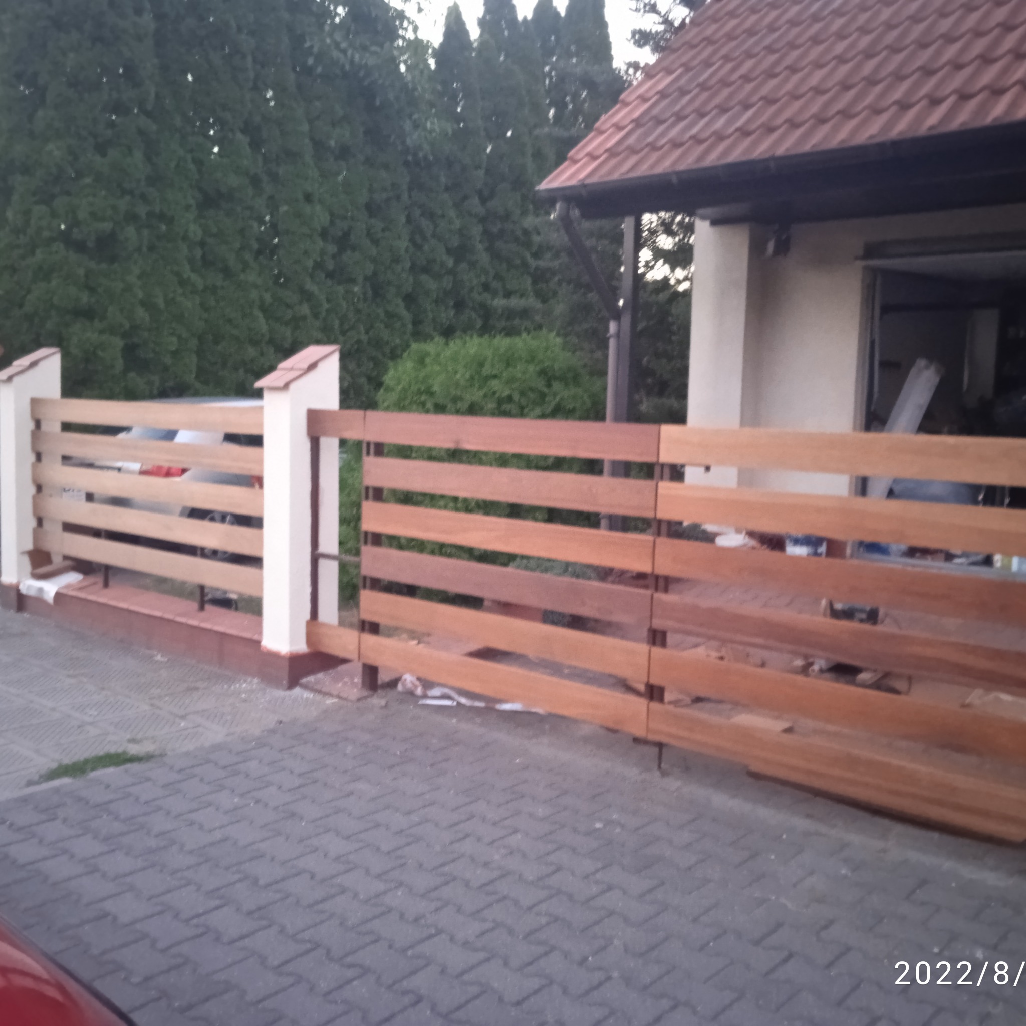 Nowoczesne, drewniane ogrodzenie posesji z poziomymi deskami w odcieniach brązu i kremowymi słupkami, widoczne z zewnątrz na tle brukowanej nawierzchni i zieleni.