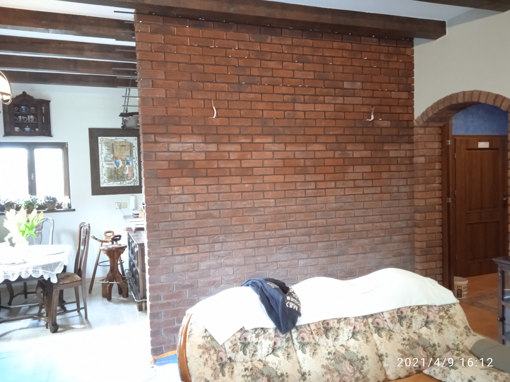 Ściana z cegły klinkierowej w salonie, widoczne drewniane belki stropowe i łukowe przejście do innego pomieszczenia. Na pierwszym planie sofa z narzutą i ubraniem.