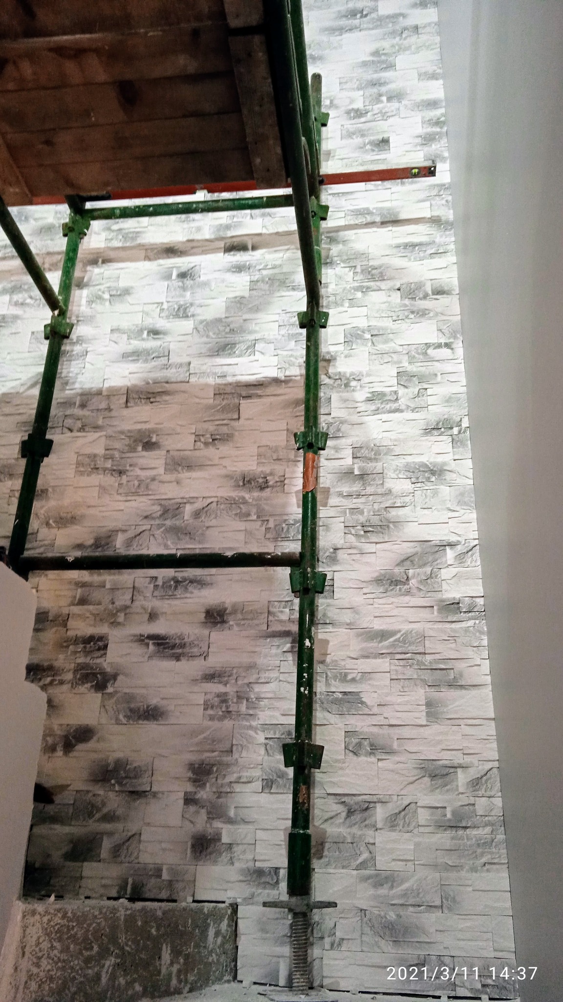 Ściana wykończona dekoracyjną, szarą cegłą, widoczna z perspektywy rusztowania w trakcie prac wykończeniowych.