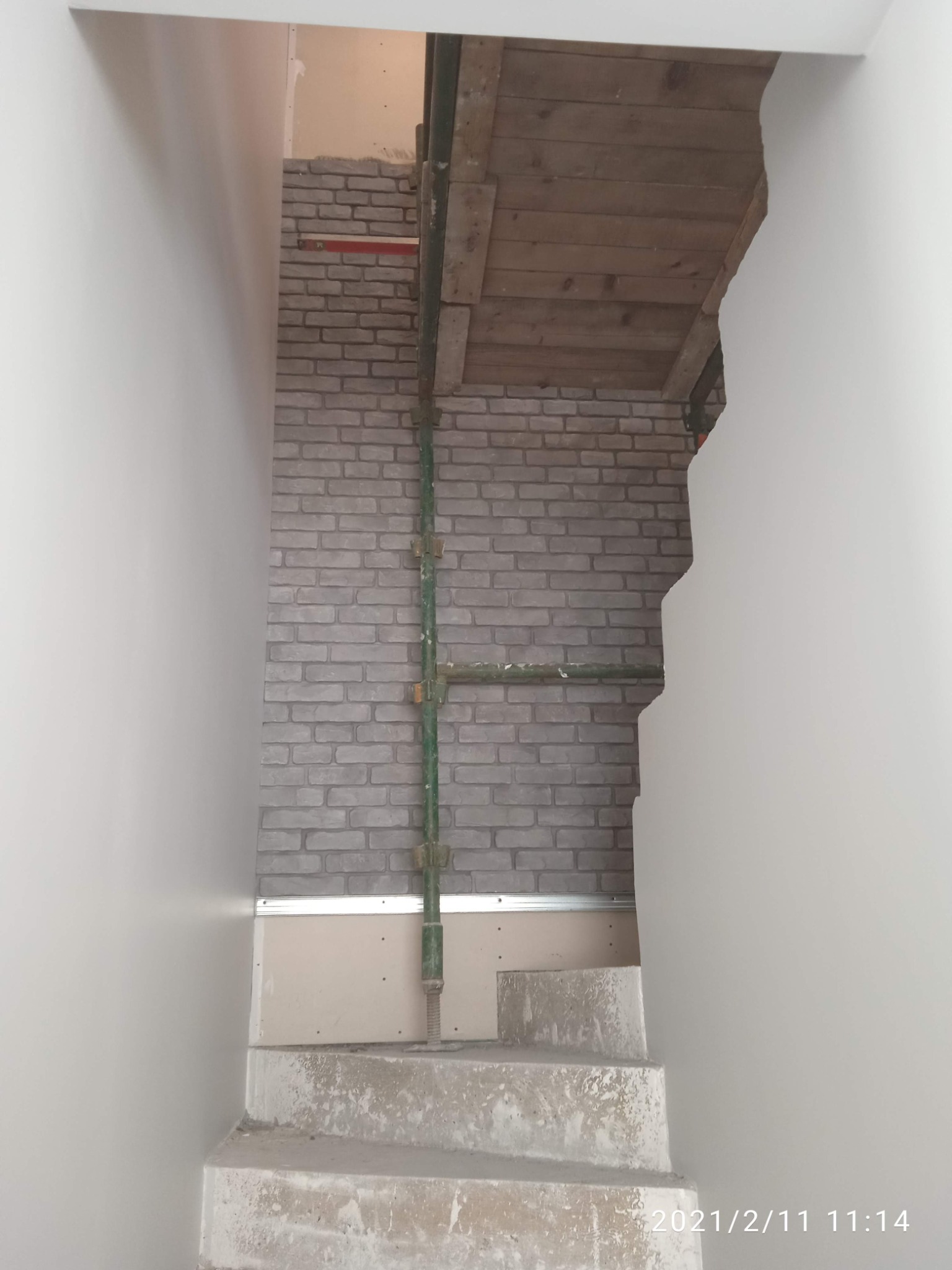 Wnętrze w trakcie prac wykończeniowych: szara cegła dekoracyjna na ścianie, częściowo zakryta rusztowaniem, widoczne płyty karton-gips i surowe schody betonowe w korytarzu.