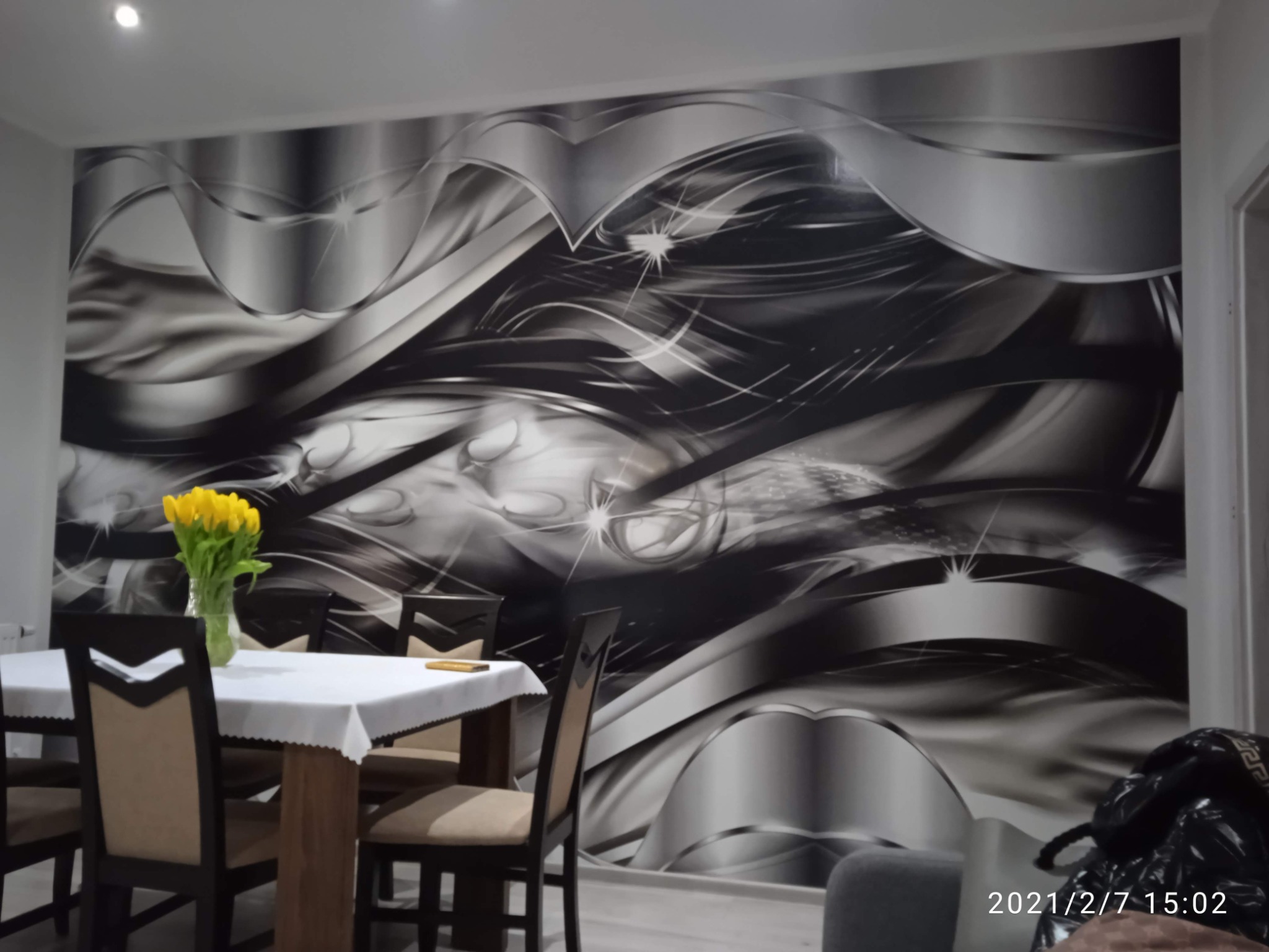 Jadalnia z fototapetą w odcieniach szarości z motywem abstrakcyjnych, metalicznych fal i refleksów świetlnych. Na pierwszym planie stół nakryty białym obrusem z bukietem żółtych tulipanów.