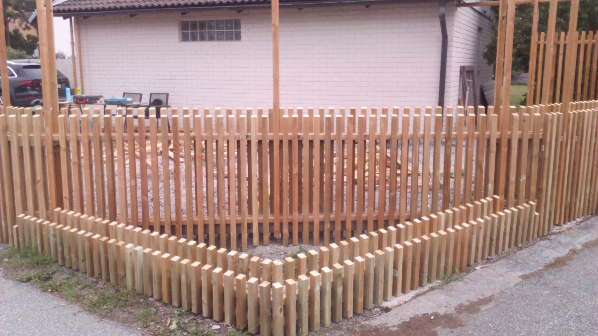 Konstrukcja drewnianego ogrodzenia z pionowych listew, częściowo ukończona, z widocznymi elementami wzdłuż krawędzi chodnika, na tle jasnego budynku z cegły.