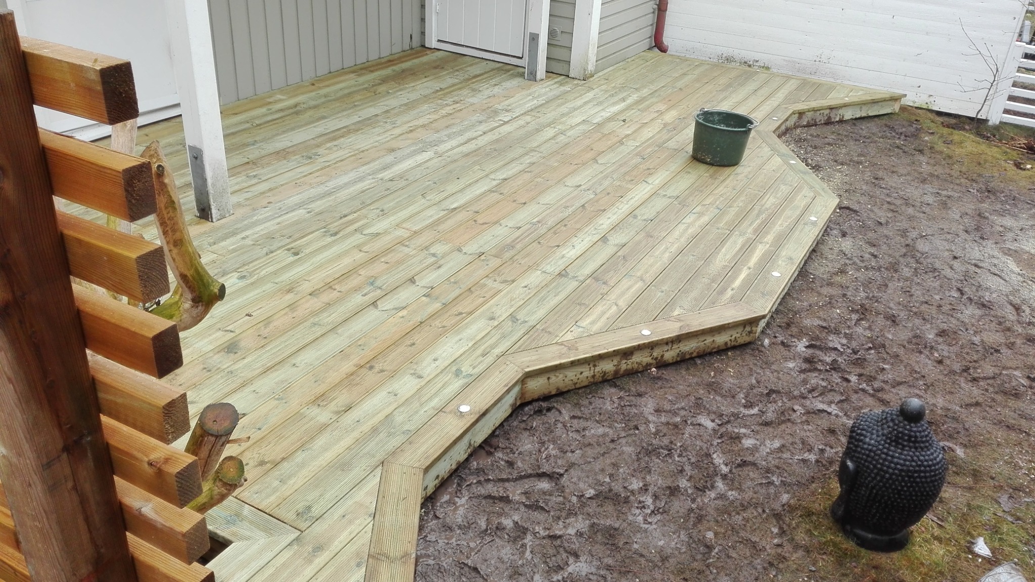 Świeżo wybudowany drewniany taras z oświetleniem punktowym, widok z boku na ziemię z błotem i dekoracyjną statuetką Buddy, w tle ściana budynku z szarymi deskami.