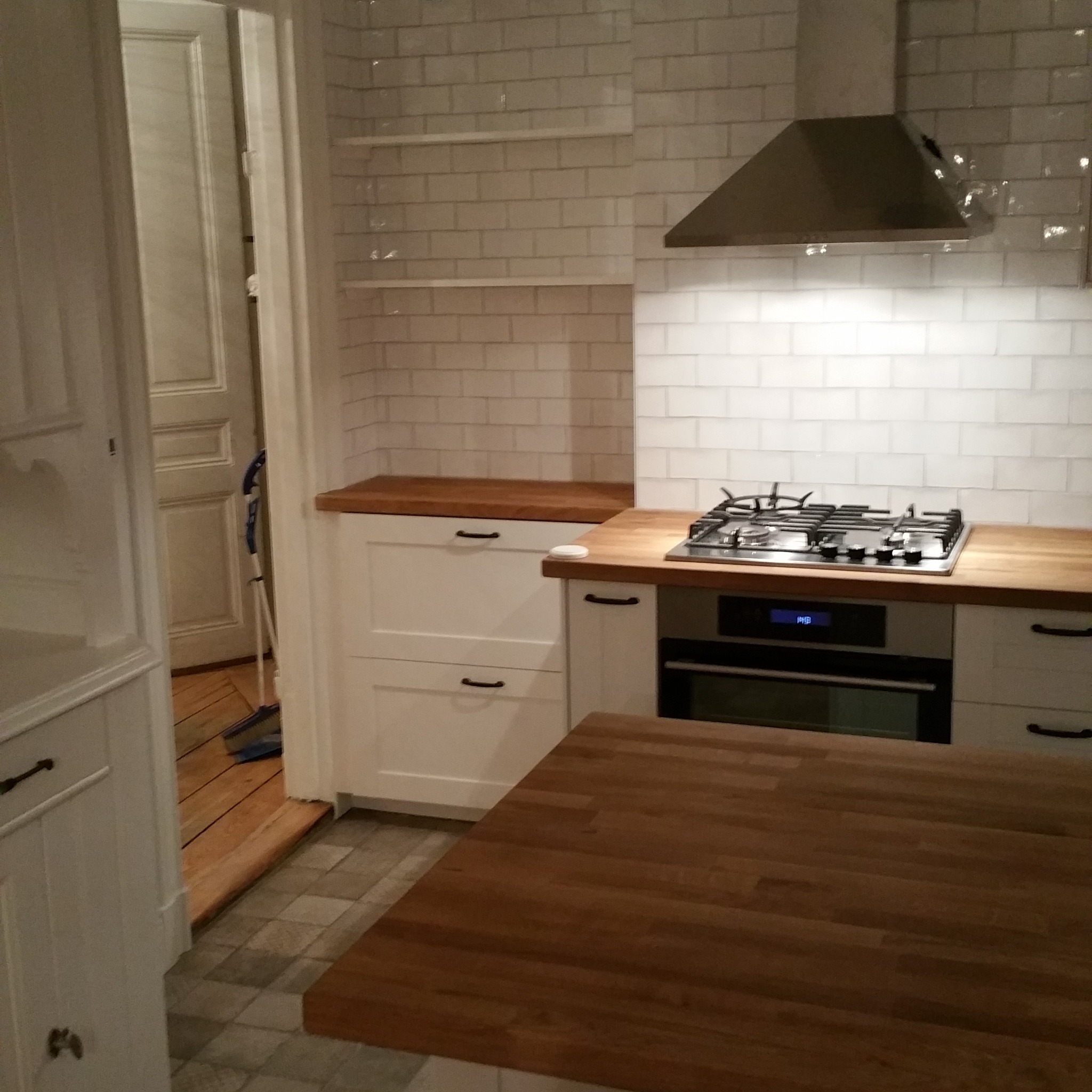 Kuchnia z białymi szafkami, drewnianym blatem i płytkami imitującymi cegłę na ścianie, widoczny fragment wyspy kuchennej z drewnianym blatem oraz otwarte drzwi do innego pomieszczenia z mopem...