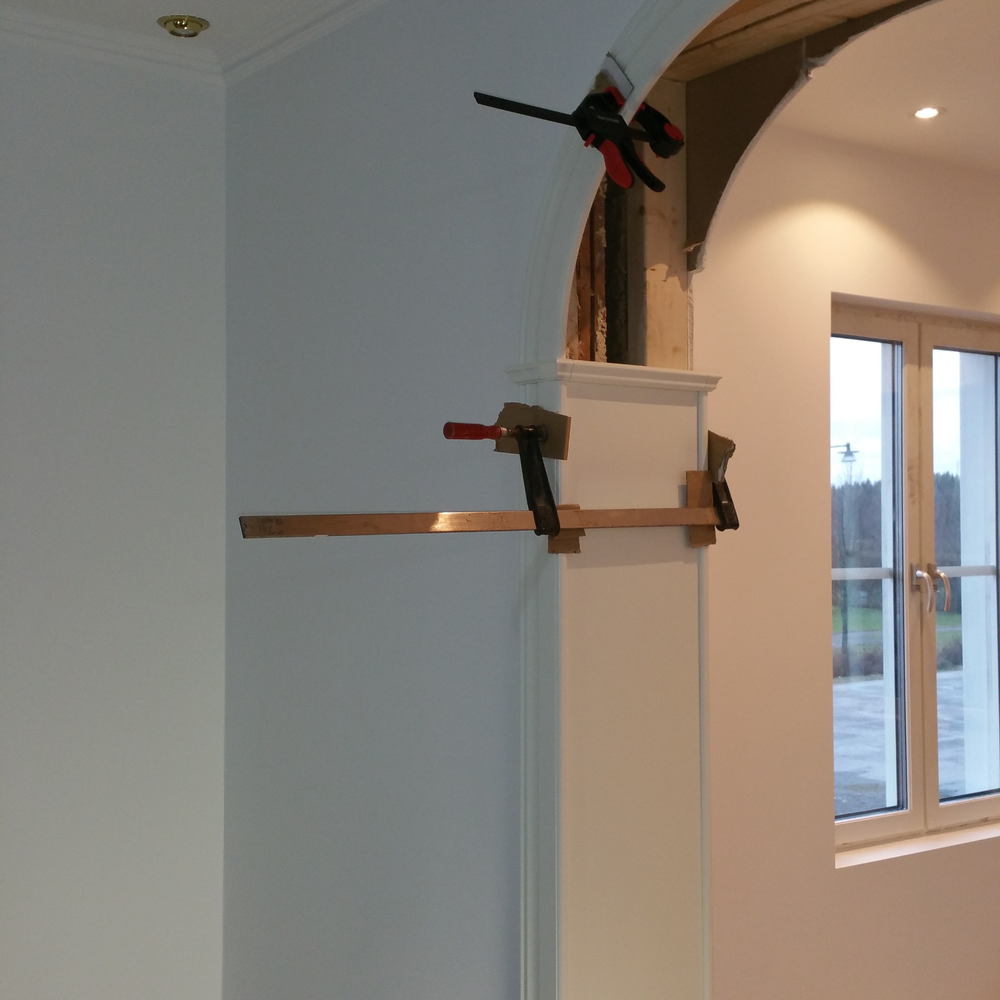 Montaż łuku drzwiowego z użyciem drewnianych rozpórek i zacisków stolarskich. Widoczne surowe wykończenie wnętrza łuku. W tle okno z widokiem na zewnątrz.