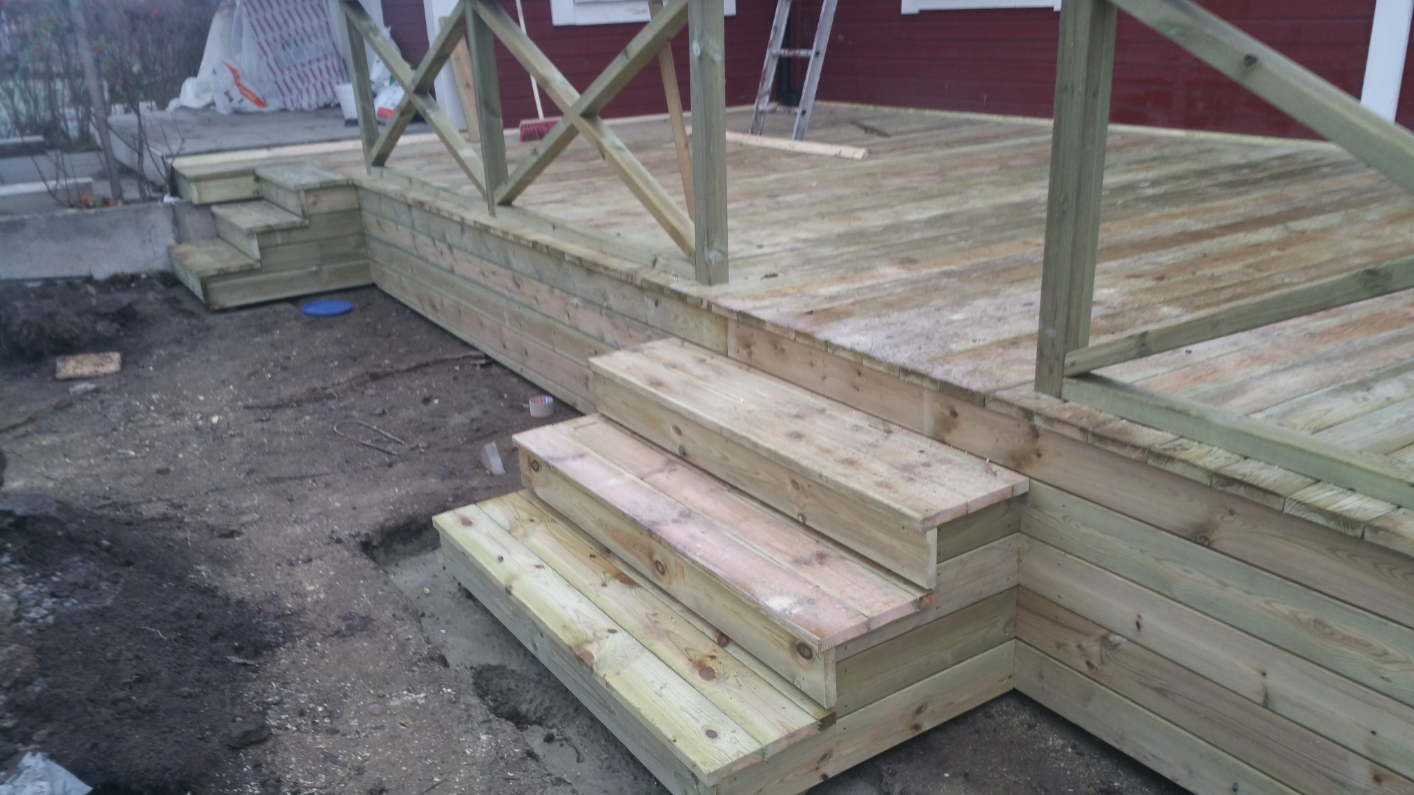 Nowo wybudowany drewniany taras z balustradą i schodami, widoczny w surowym stanie przed wykończeniem, z ziemią wokół i fragmentem czerwonego budynku w tle.