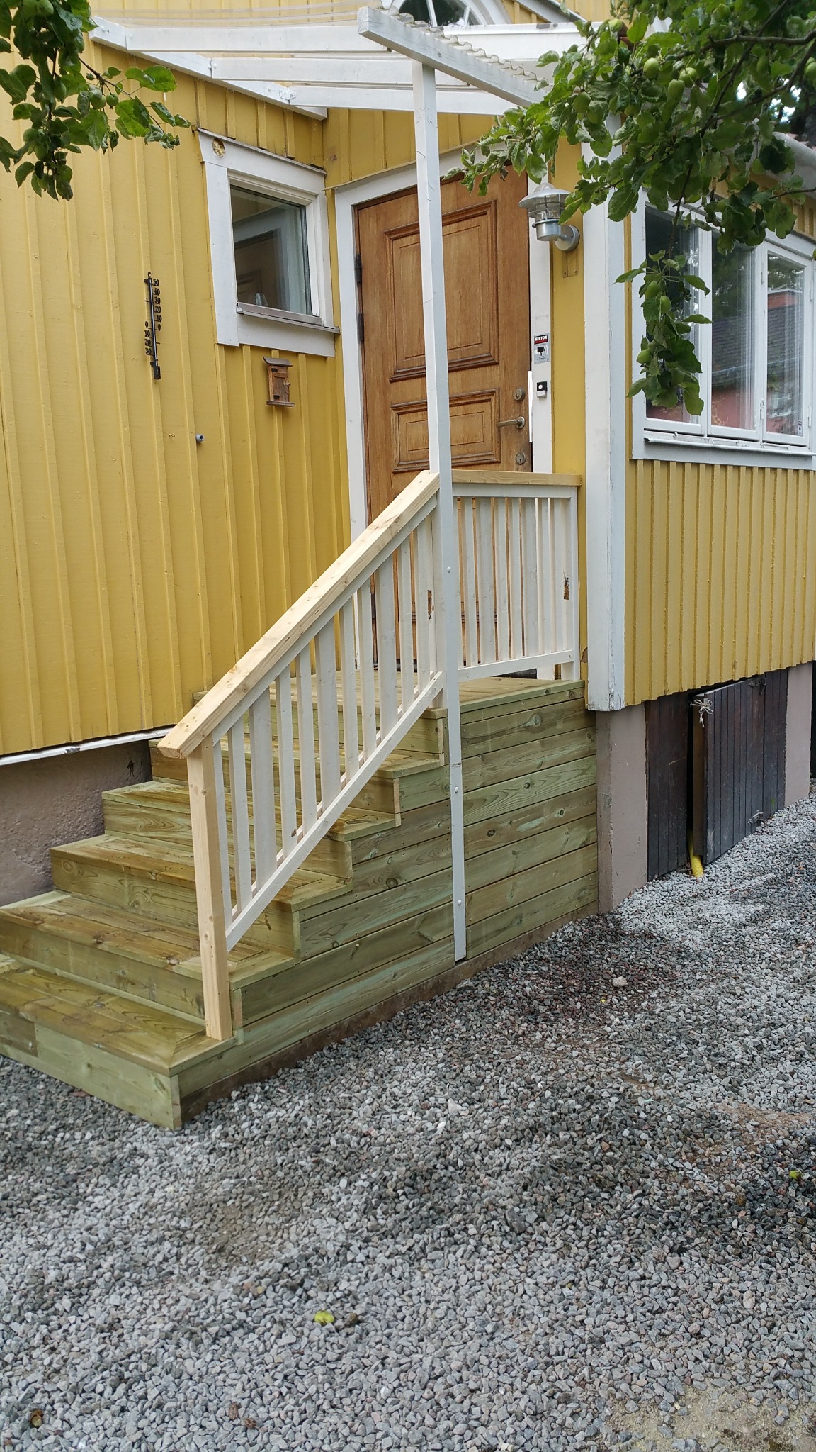 Nowe drewniane schody z białą balustradą prowadzące do żółtego domu z drewnianą elewacją i brązowymi drzwiami wejściowymi.