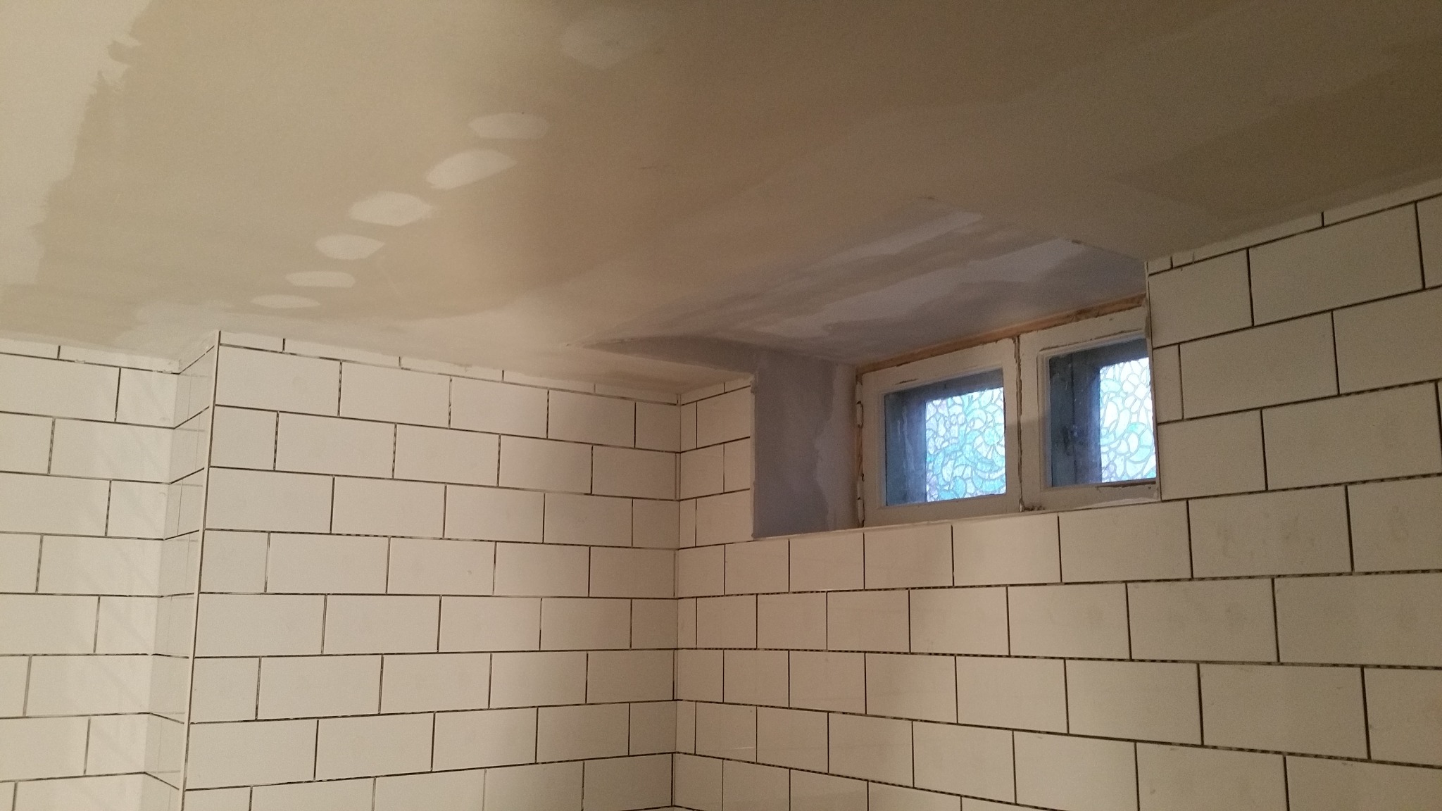 Ściana wyłożona białymi, prostokątnymi płytkami z ciemną fugą, sufit w trakcie szpachlowania z widocznymi łatami gładzi, okno z ozdobną szybą w górnej części ściany.