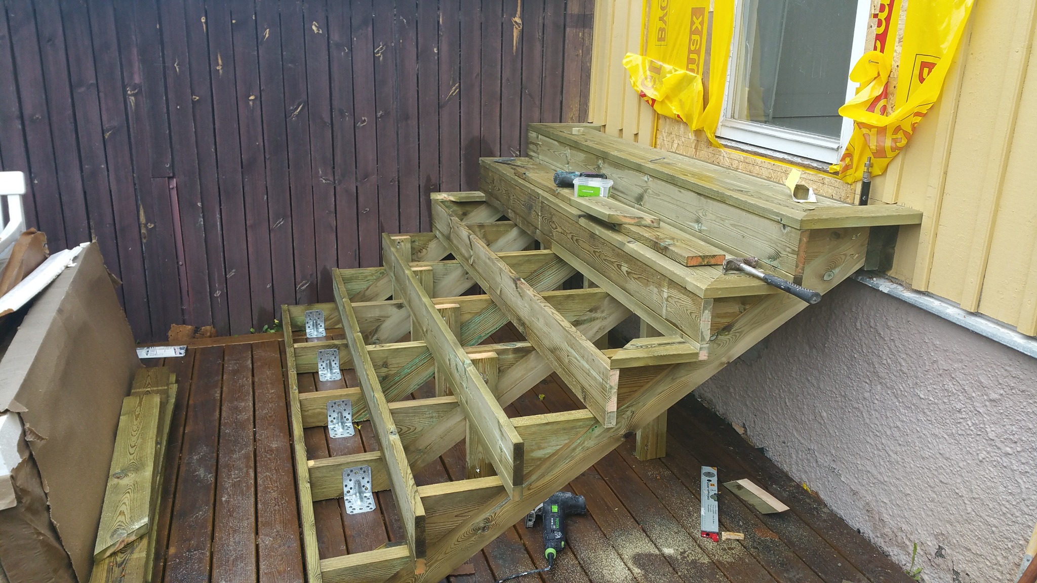 Konstrukcja drewnianych schodów zewnętrznych w trakcie montażu, widoczne elementy mocujące, narzędzia i deski na tarasie z brązowych desek.