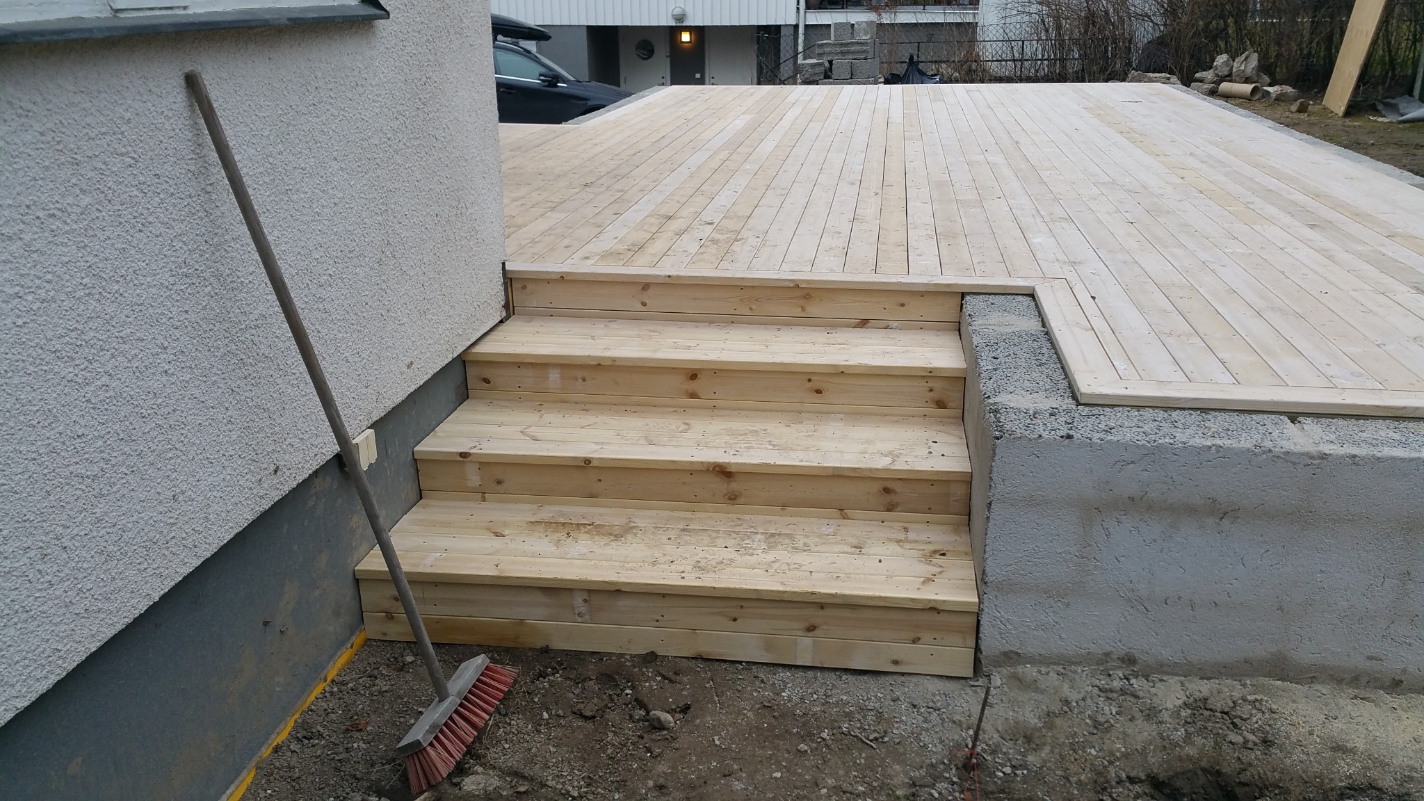 Nowe drewniane schody prowadzące na taras z jasnych desek, opierające się o szarą ścianę budynku, obok stoi czerwona zmiotka.