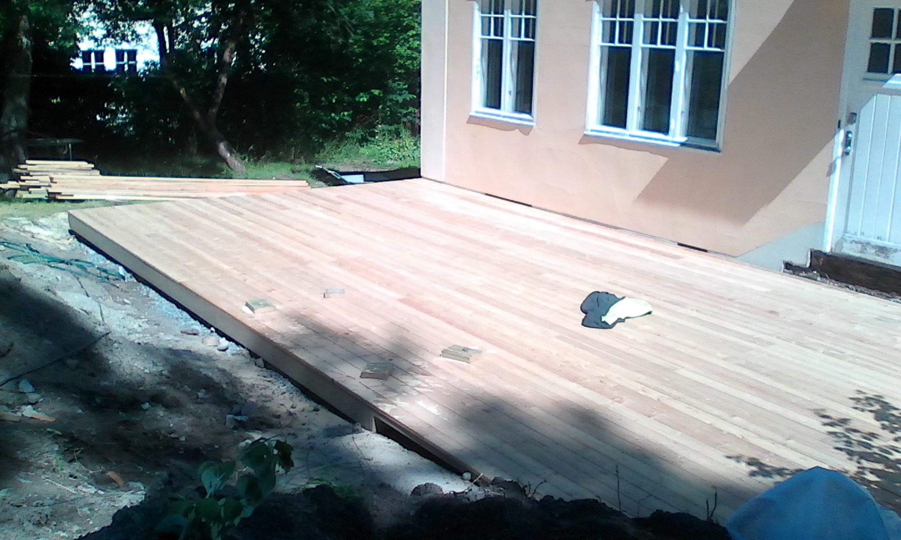 Nowy drewniany taras przylegający do domu z beżową elewacją i białymi oknami, widoczne deski tarasowe ułożone równolegle, w tle stos desek i zieleń.