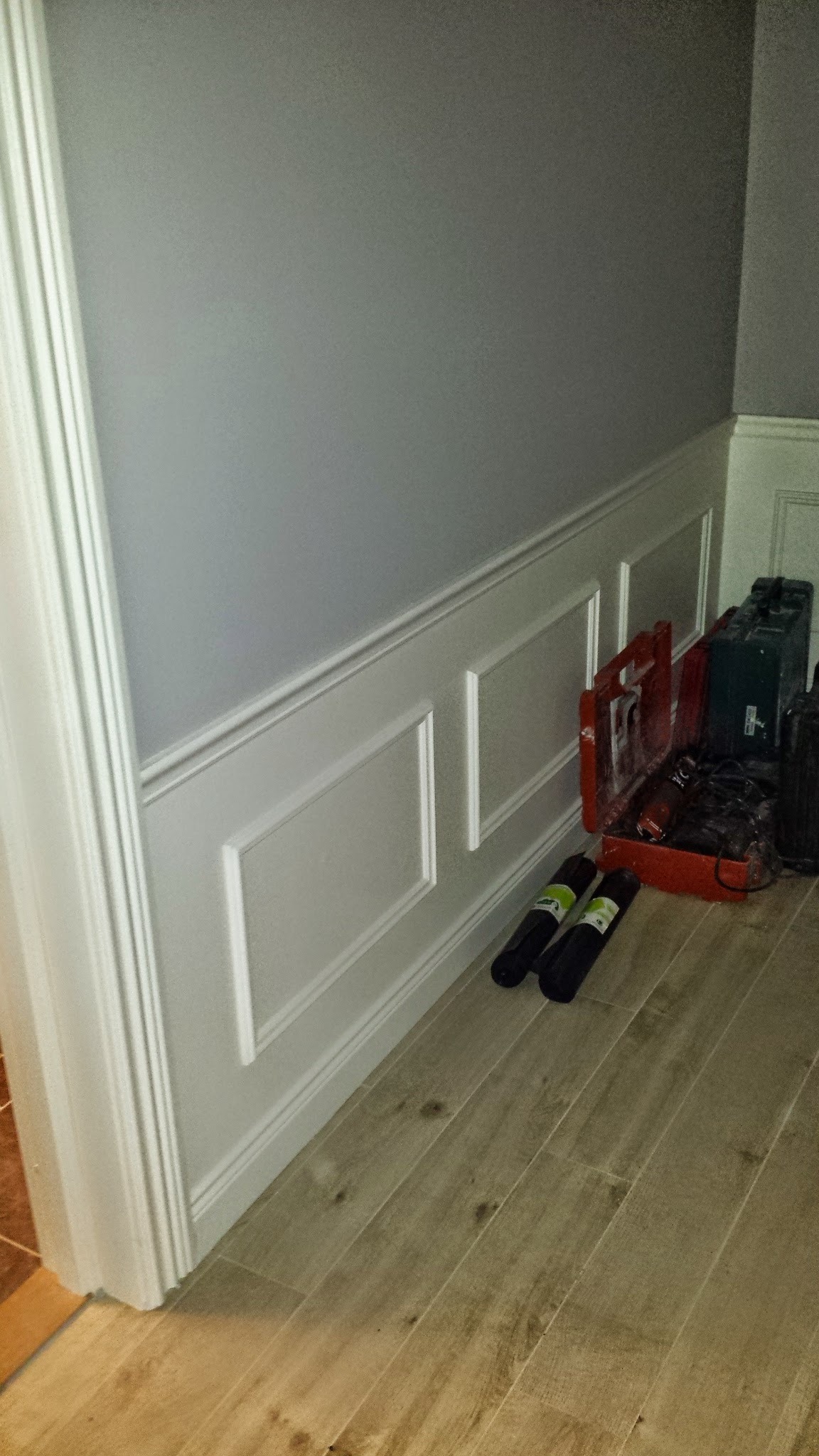 Szara ściana z dekoracyjną białą boazerią panelową, narzędzia i skrzynki narzędziowe leżące na podłodze z paneli imitujących drewno, widok z dołu.