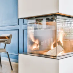Nowoczesny, przeszklony wkład kominkowy narożny z paleniskiem, w tle błękitna ściana z dekoracyjnymi panelami i fragment jadalni z drewnianym stołem.