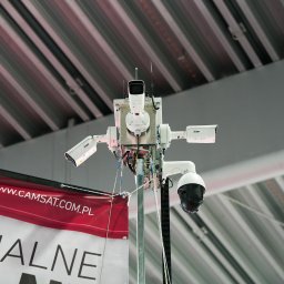 Safelock Jerzy Ziobro - Zestaw kamer przemysłowych różnego typu (obrotowa, kopułkowa, tubowa) zamontowany na metalowym słupie, widoczny baner reklamowy z adresem strony internetowej, tło stanowi sufit hali z widocznymi...