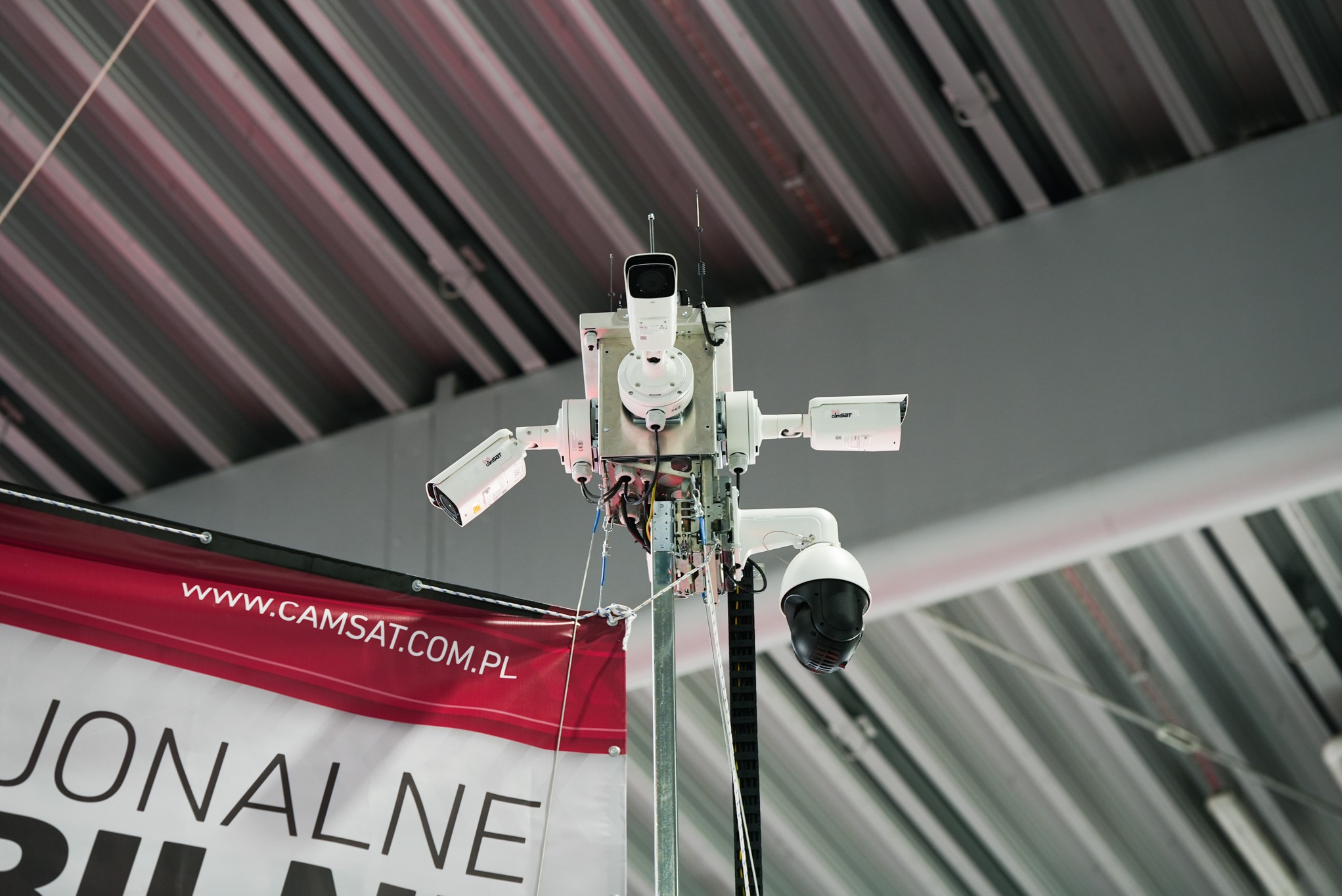 Zestaw kamer przemysłowych różnego typu (obrotowa, kopułkowa, tubowa) zamontowany na metalowym słupie, widoczny baner reklamowy z adresem strony internetowej, tło stanowi sufit hali z widocznymi...