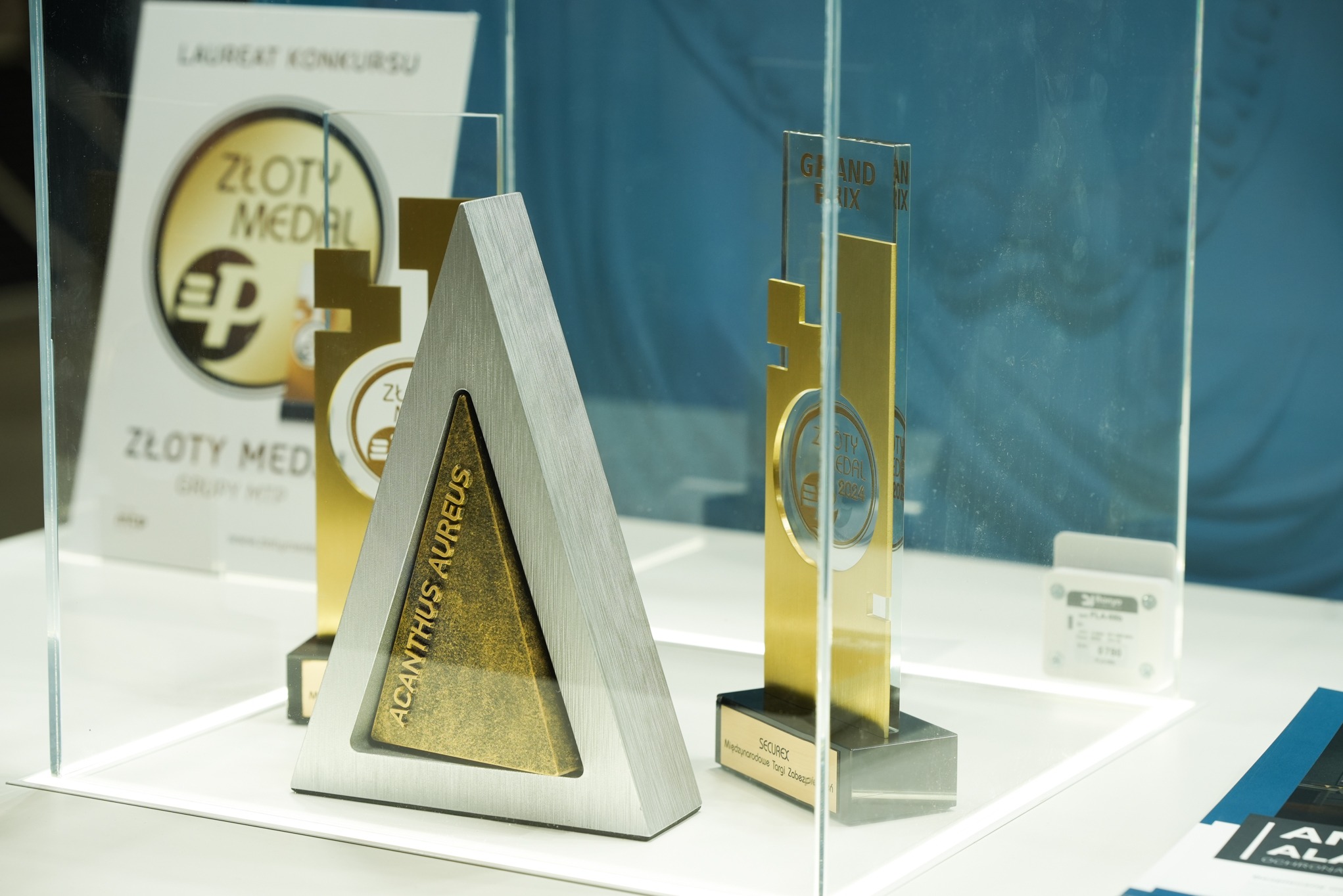 Nagrody i wyróżnienia, w tym statuetka w kształcie trójkąta z napisem 'Acanthus Aureus', prezentowane na targach Securex w Tomaszowie, prawdopodobnie związane z branżą zabezpieczeń.