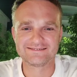 Portret uśmiechniętego mężczyzny w białej koszulce na tle zieleni i budynku, prawdopodobnie właściciel firmy remontowej.