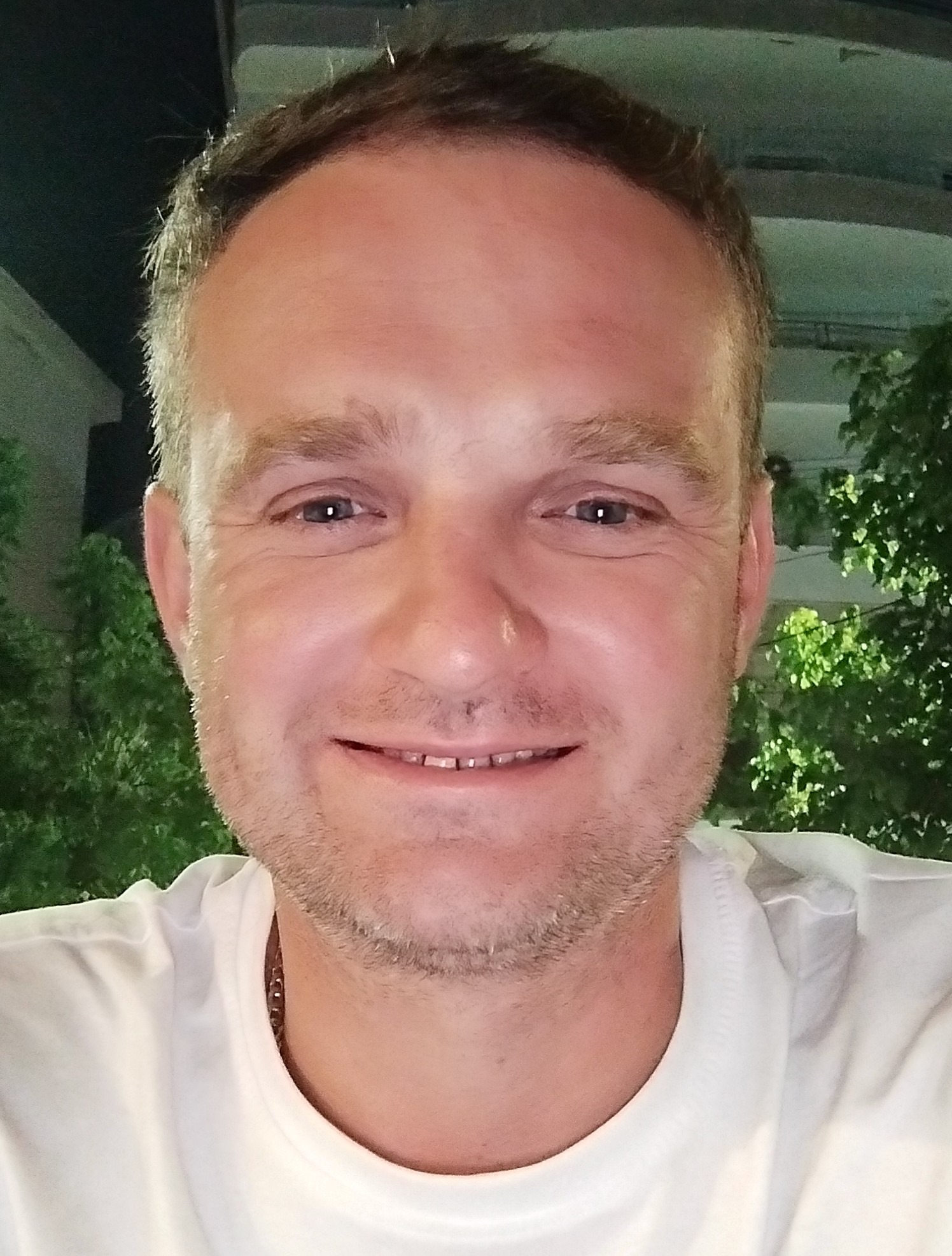 Portret uśmiechniętego mężczyzny w białej koszulce na tle zieleni i budynku, prawdopodobnie właściciel firmy remontowej.