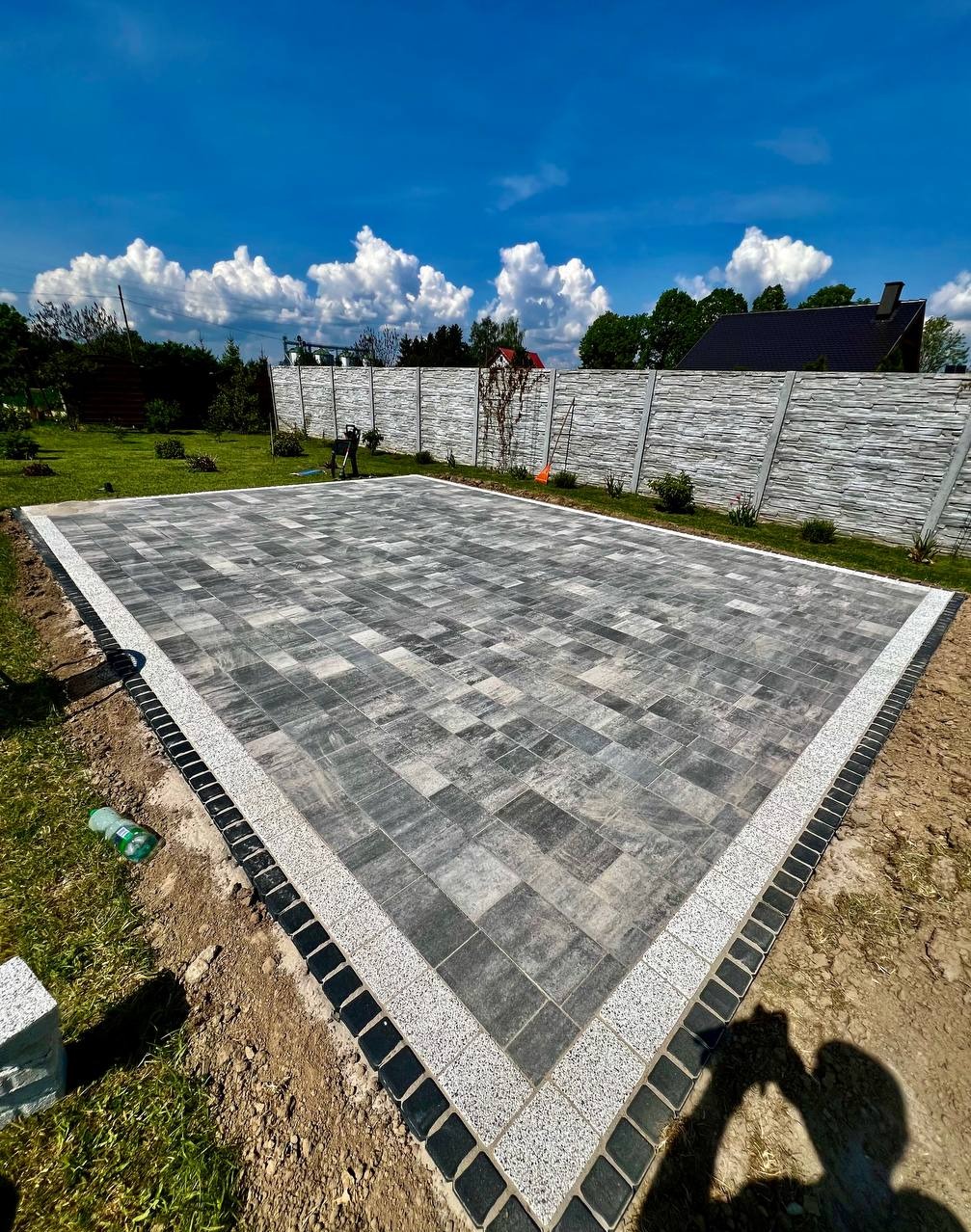 Nowo ułożona, prostokątna nawierzchnia z szarej kostki brukowej o nieregularnym wzorze, obramowana jasnym i ciemnym pasem kostki, widok z góry na tle zielonego trawnika i betonowego ogrodzenia.